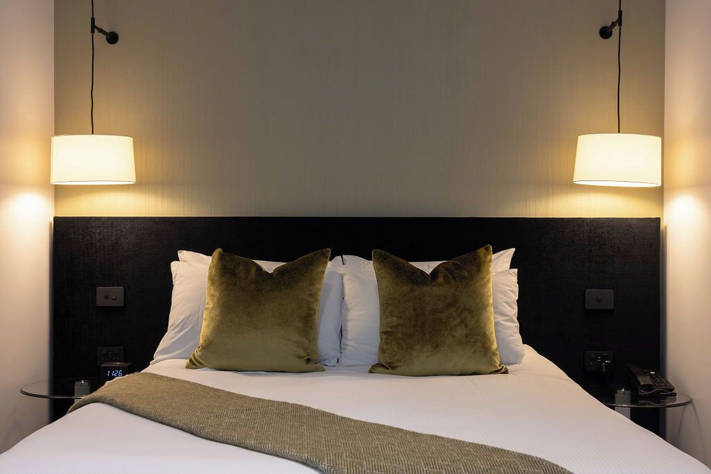 A hotel room with a large bed, two bedside lamps, a nightstand with a phone, and a wall clock.