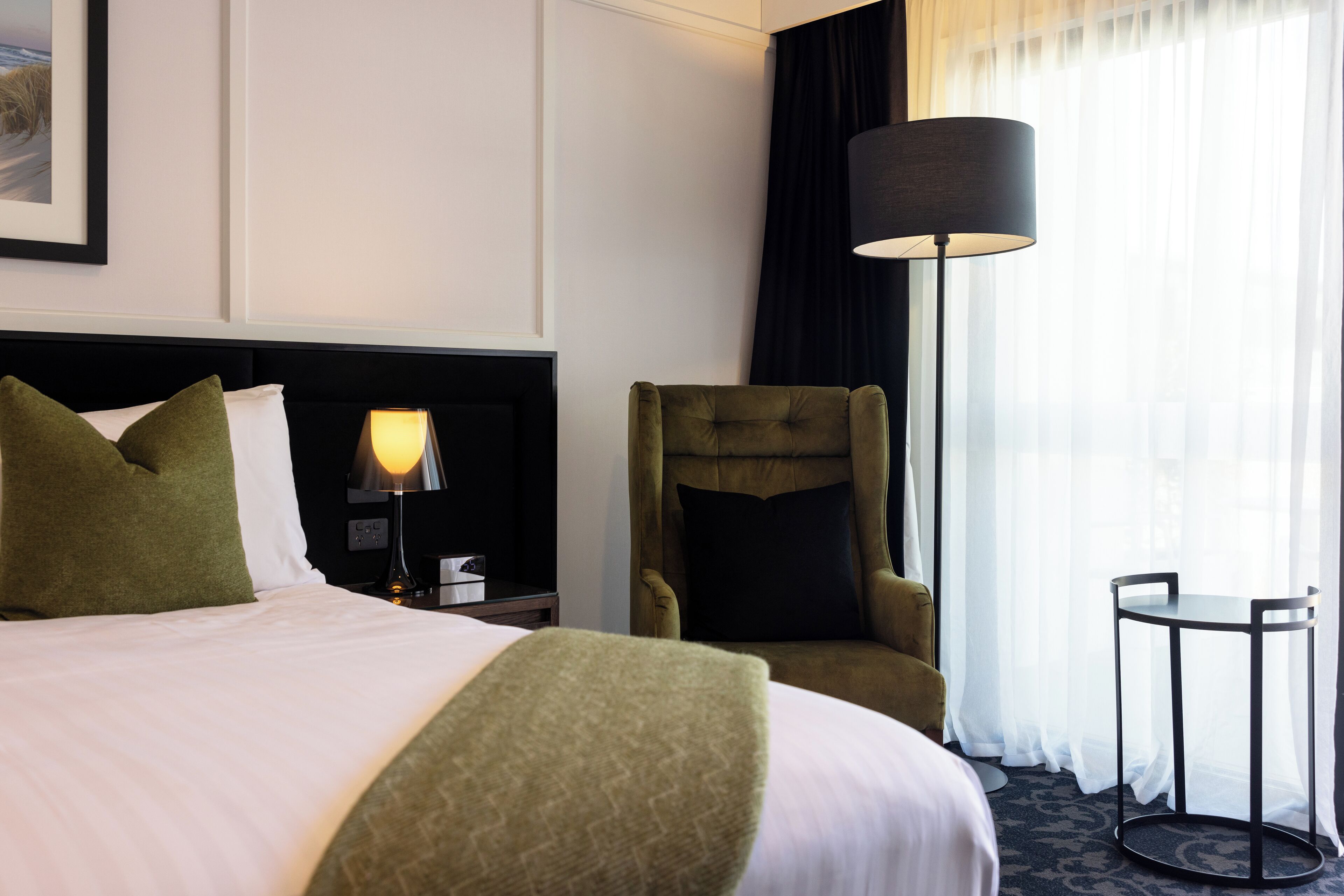 A hotel room with a large bed, a green armchair, a small table, a lamp, and a window with sheer curtains.