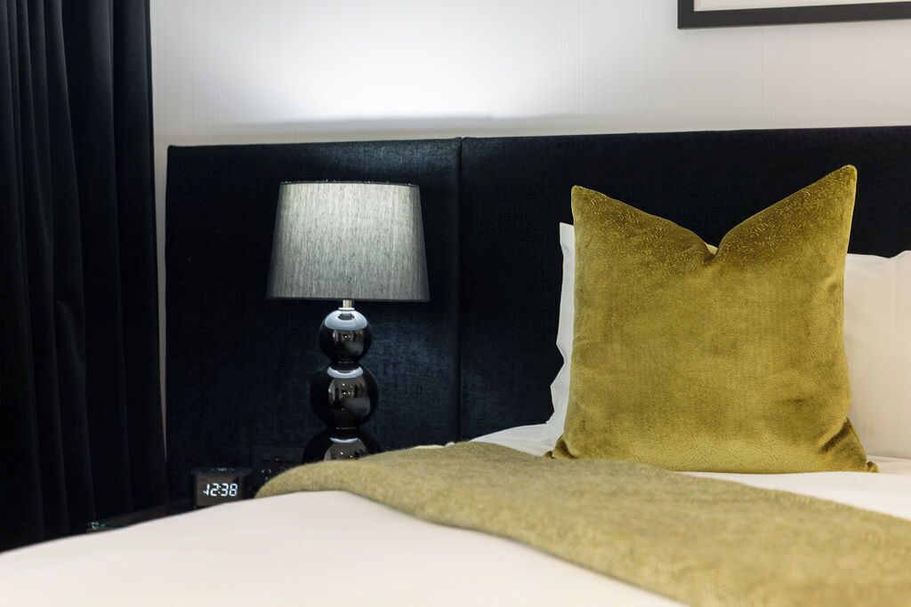 Una cama bien hecha con una manta dorada, un cabecero negro, una lámpara de noche y un cojín dorado.
