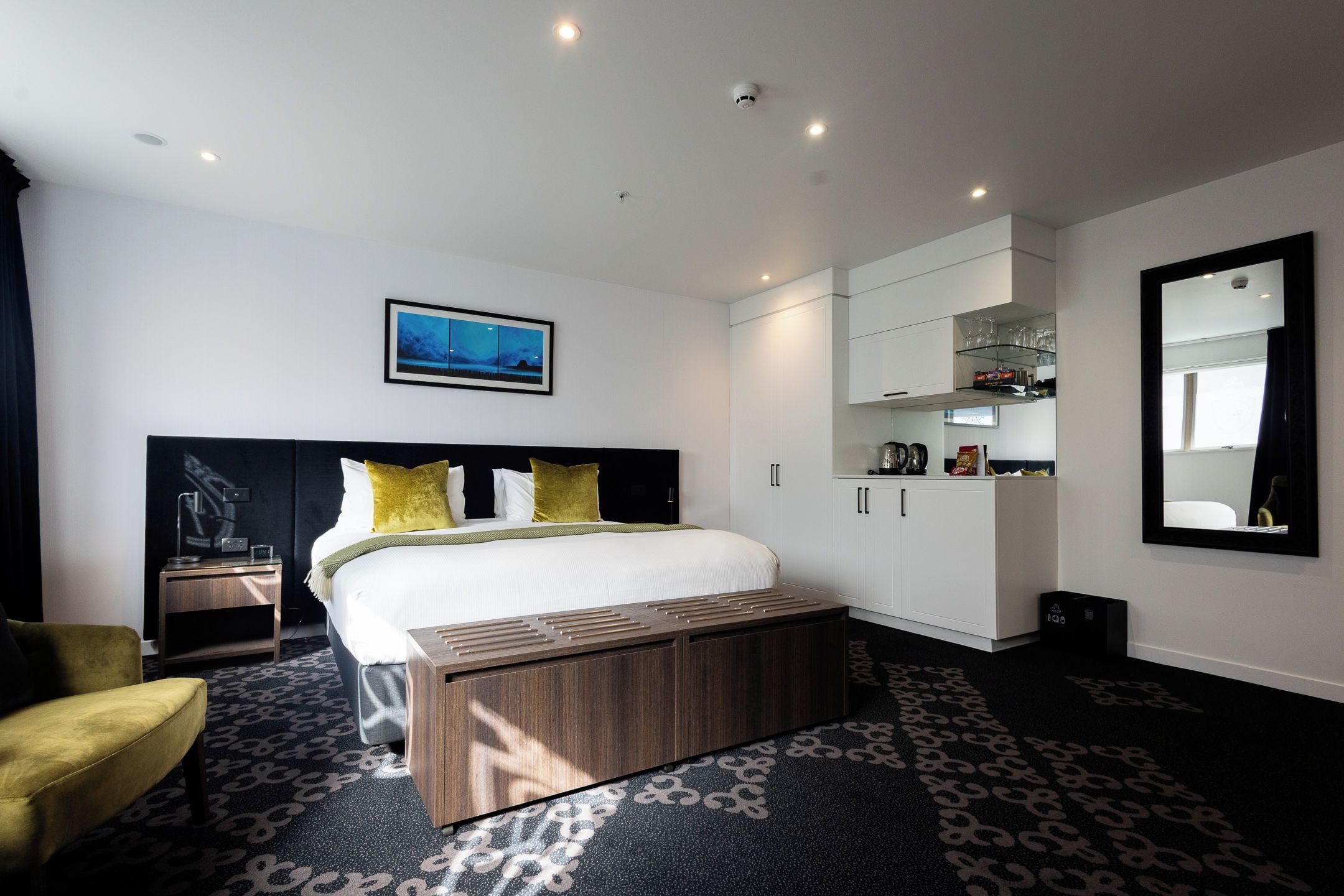 A modern hotel room with a large bed, a bedside table, a bench, a wardrobe, and a mirror.