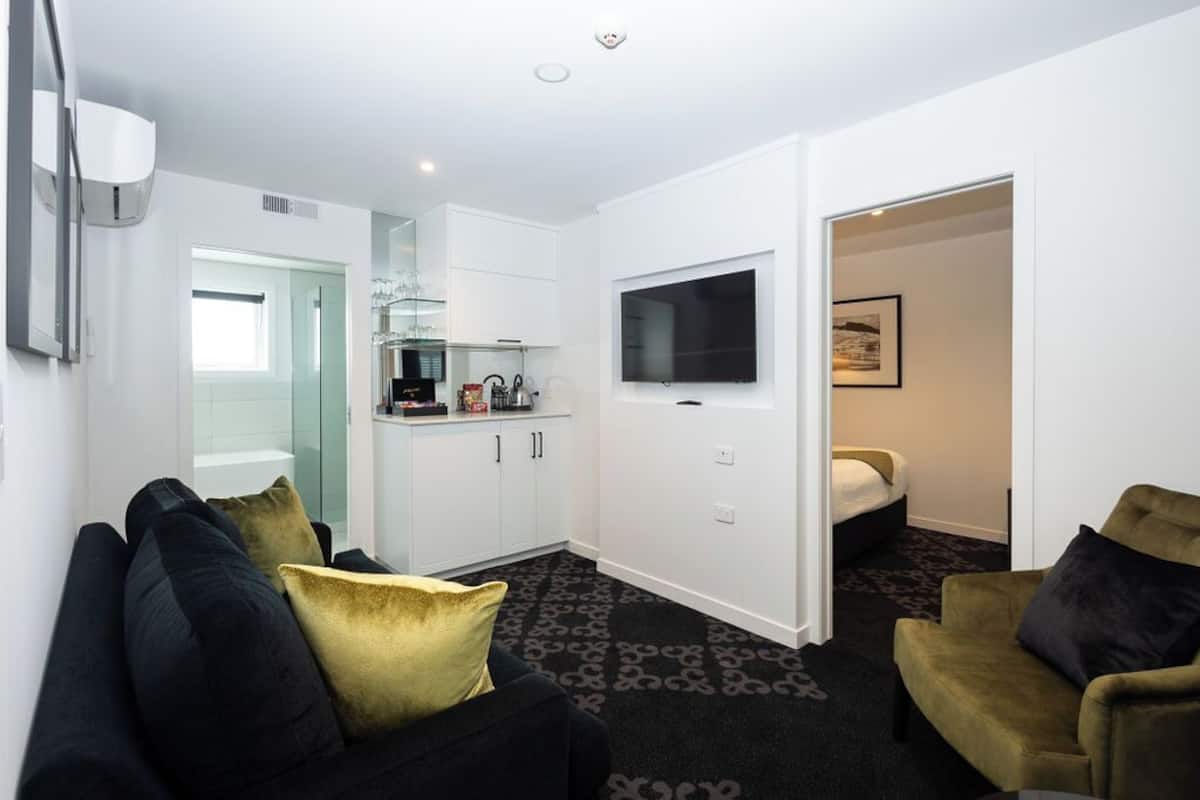 A compact hotel room with a kitchenette, a flat-screen TV mounted on the wall, a bed, and a seating area with two sofas.