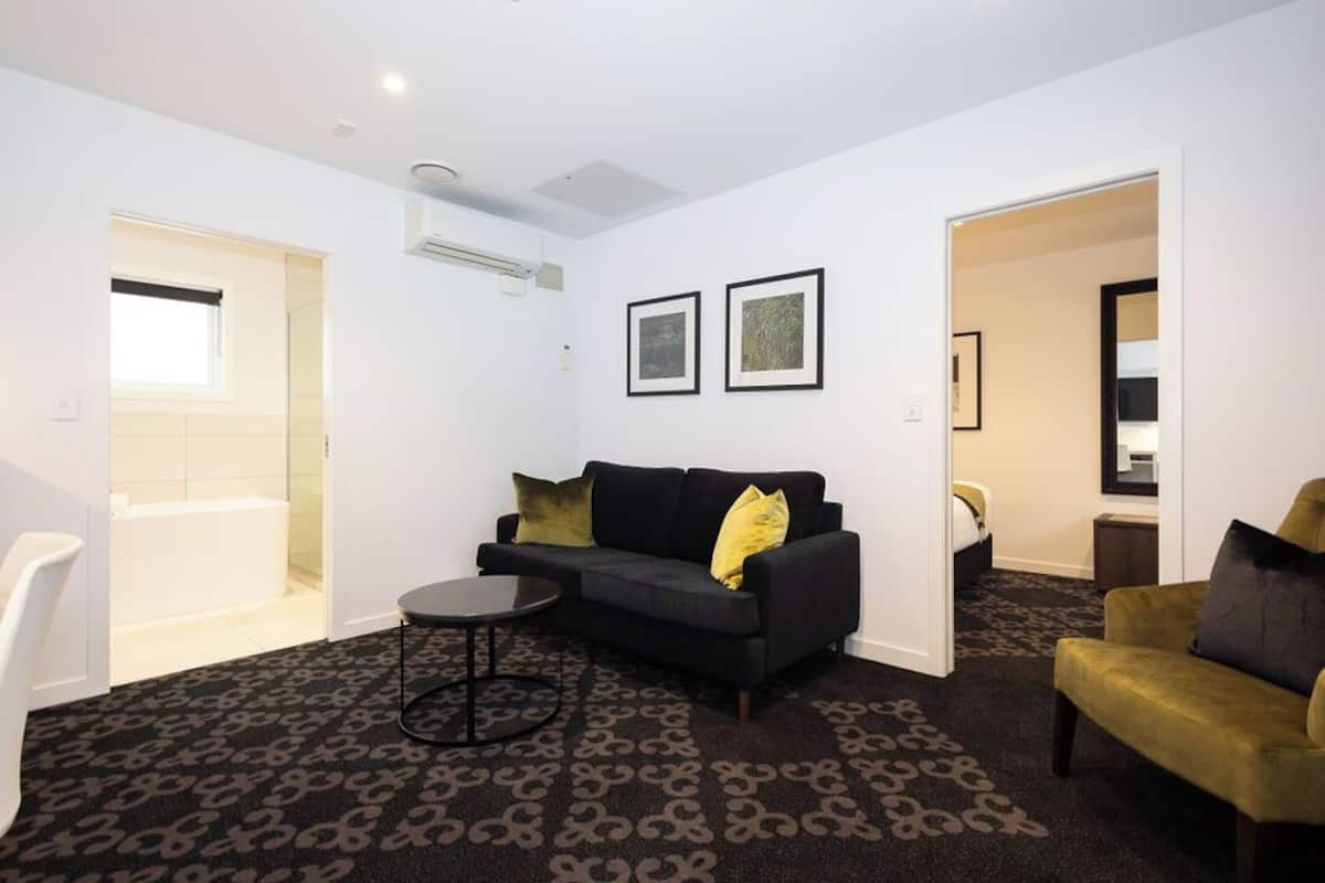 A modern living room with a black sofa, a round coffee table, and a patterned carpet. There are two framed pictures on the wall, a window, and a door leading to a bathroom.