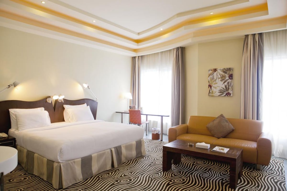 Quarto de hotel com uma cama grande, um sofá, uma mesa de centro e uma escrivaninha com luminária.