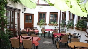 Un patio exterior con sillas y mesas de mimbre, rodeado de paredes blancas y ventanas con plantas.