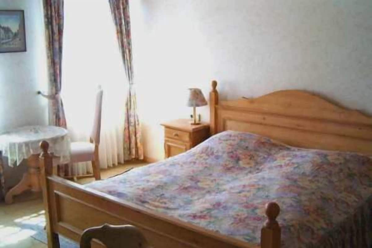 Ein Schlafzimmer mit einem hölzernen Bett, einem Nachttisch mit Lampe, einem Sessel und einem Fenster mit Vorhängen.