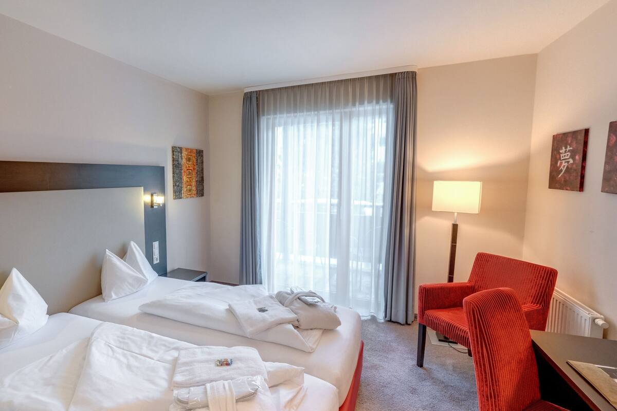 A hotel room with two beds, a red chair, a desk, a lamp, and a window with curtains.