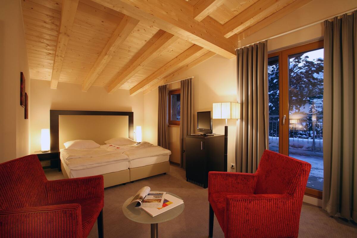 A hotel room with a wooden ceiling, a bed, a TV, a desk, and two red chairs.