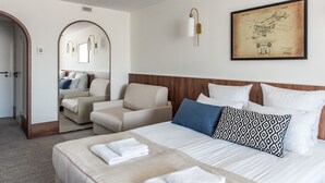 Habitación de hotel con cama, zona de estar con sillón, espejo grande y un cuadro en la pared.