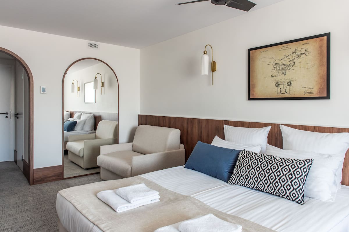 A hotel room with a bed, a sitting area with a chair, a large mirror, and a framed picture on the wall.