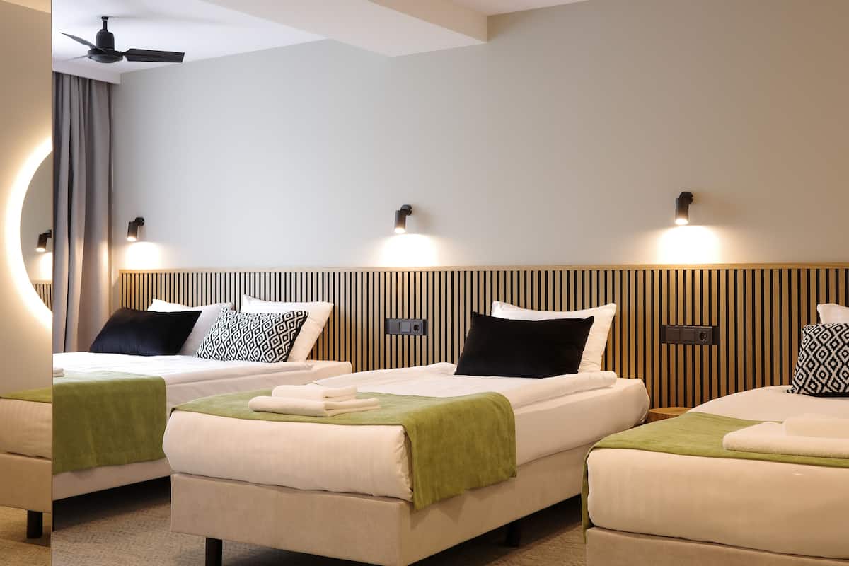 A hotel room with three beds, a ceiling fan, wall-mounted lights, and a wooden panel design on the wall.