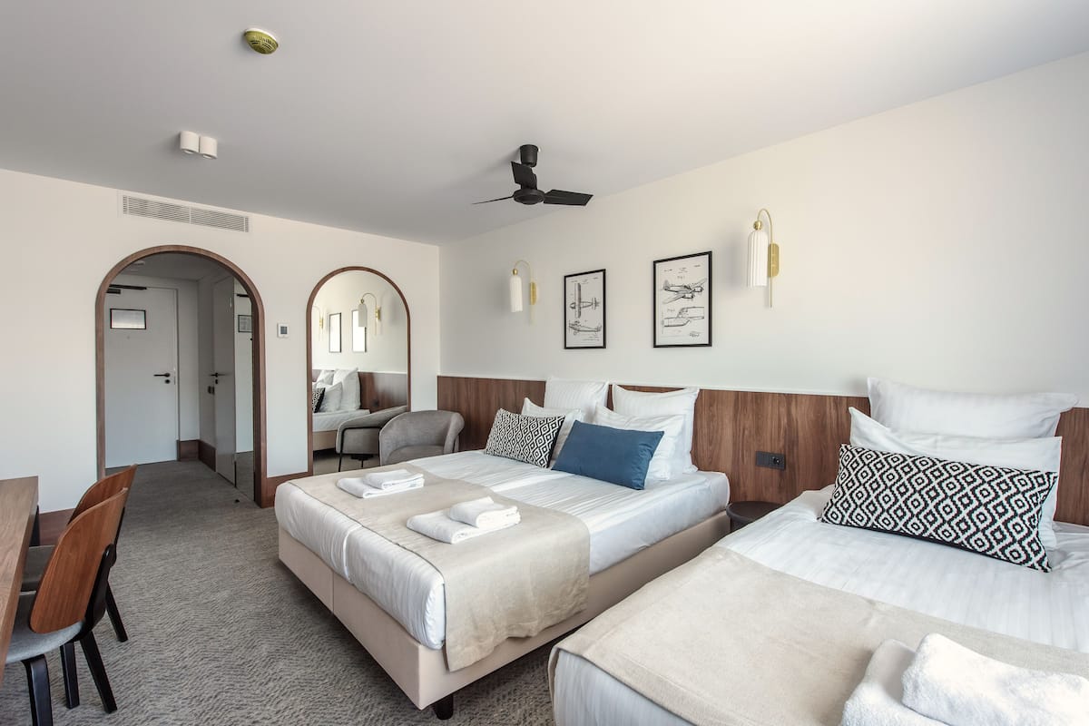 A hotel room with two beds, a desk, and a chair. There are framed pictures on the wall, a ceiling fan, and a light fixture.