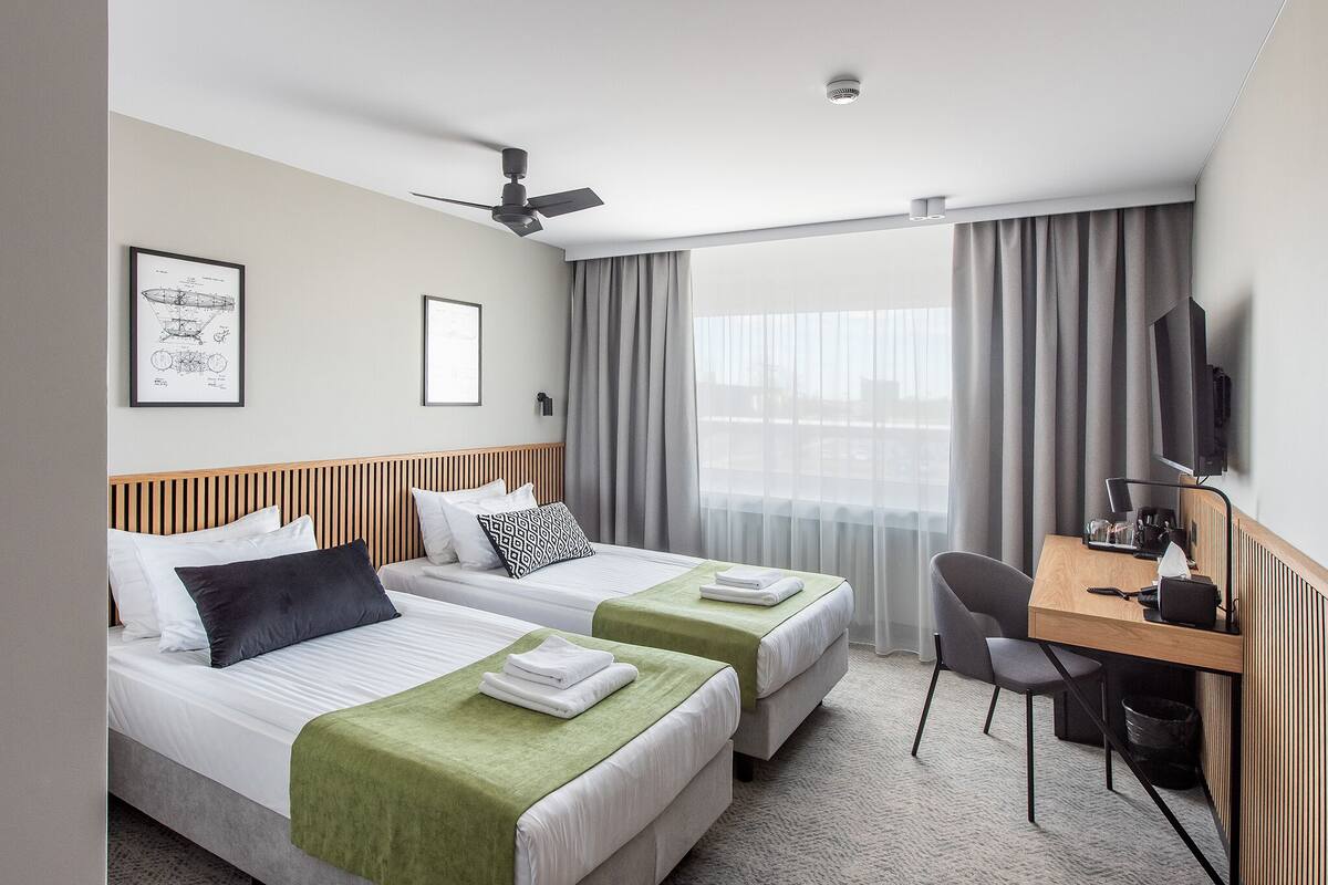 A hotel room with two beds, a desk, a chair, a ceiling fan, and a large window with curtains.