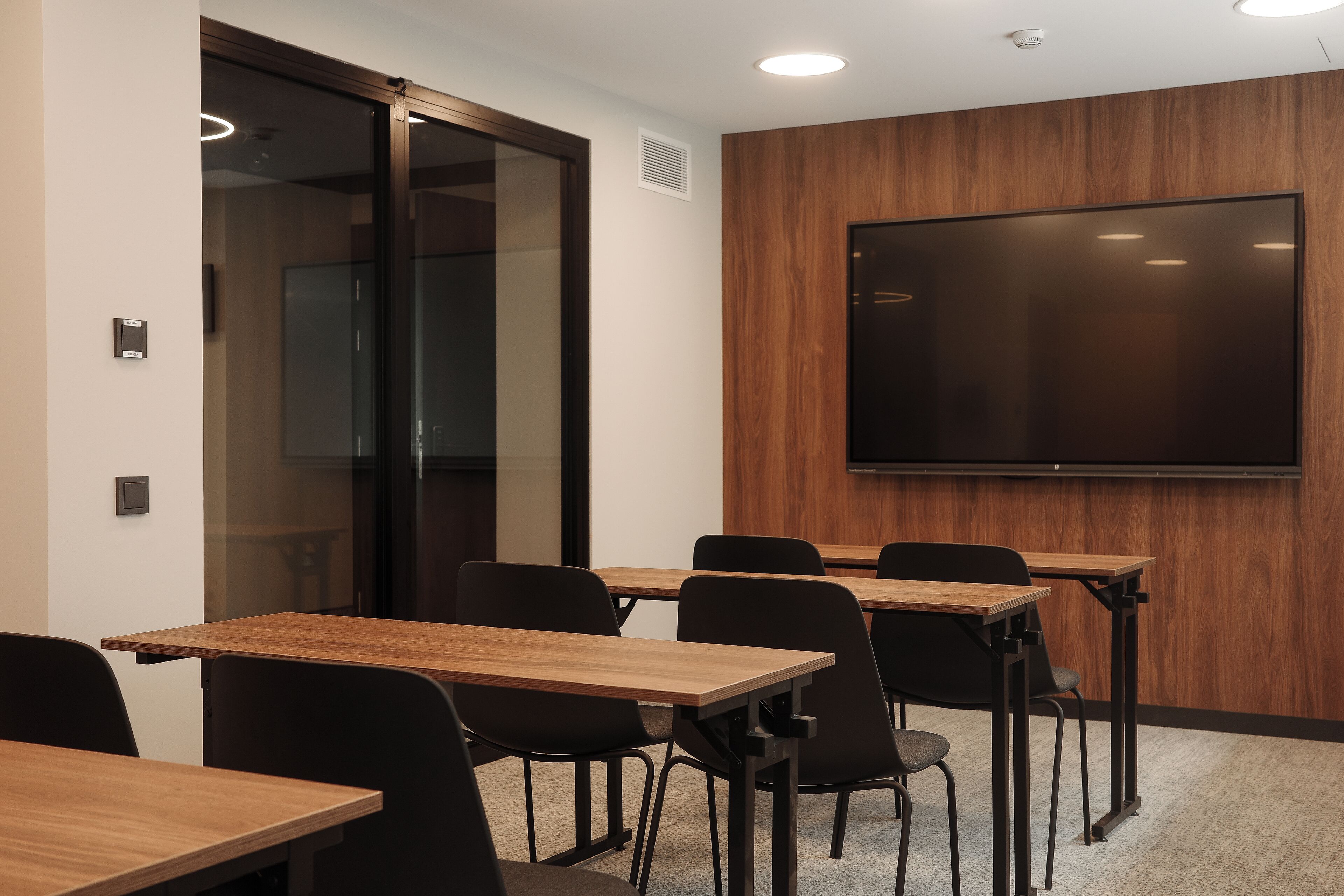 A conference room with wooden tables and black chairs, a large flat-screen TV on the wall, and a glass door leading to another room.