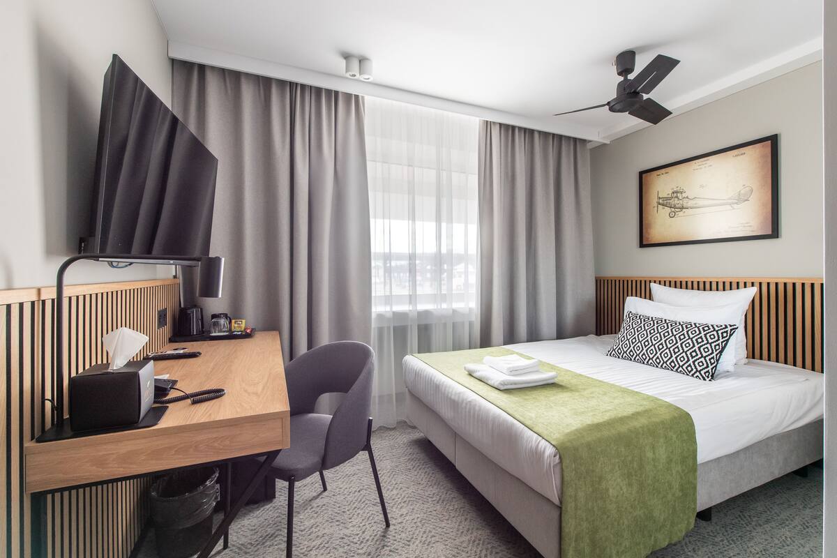 A hotel room with a bed, a desk with a computer, a chair, a ceiling fan, and a window with curtains.