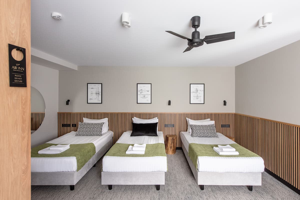 A hotel room with three single beds, a ceiling fan, and framed architectural drawings on the wall.