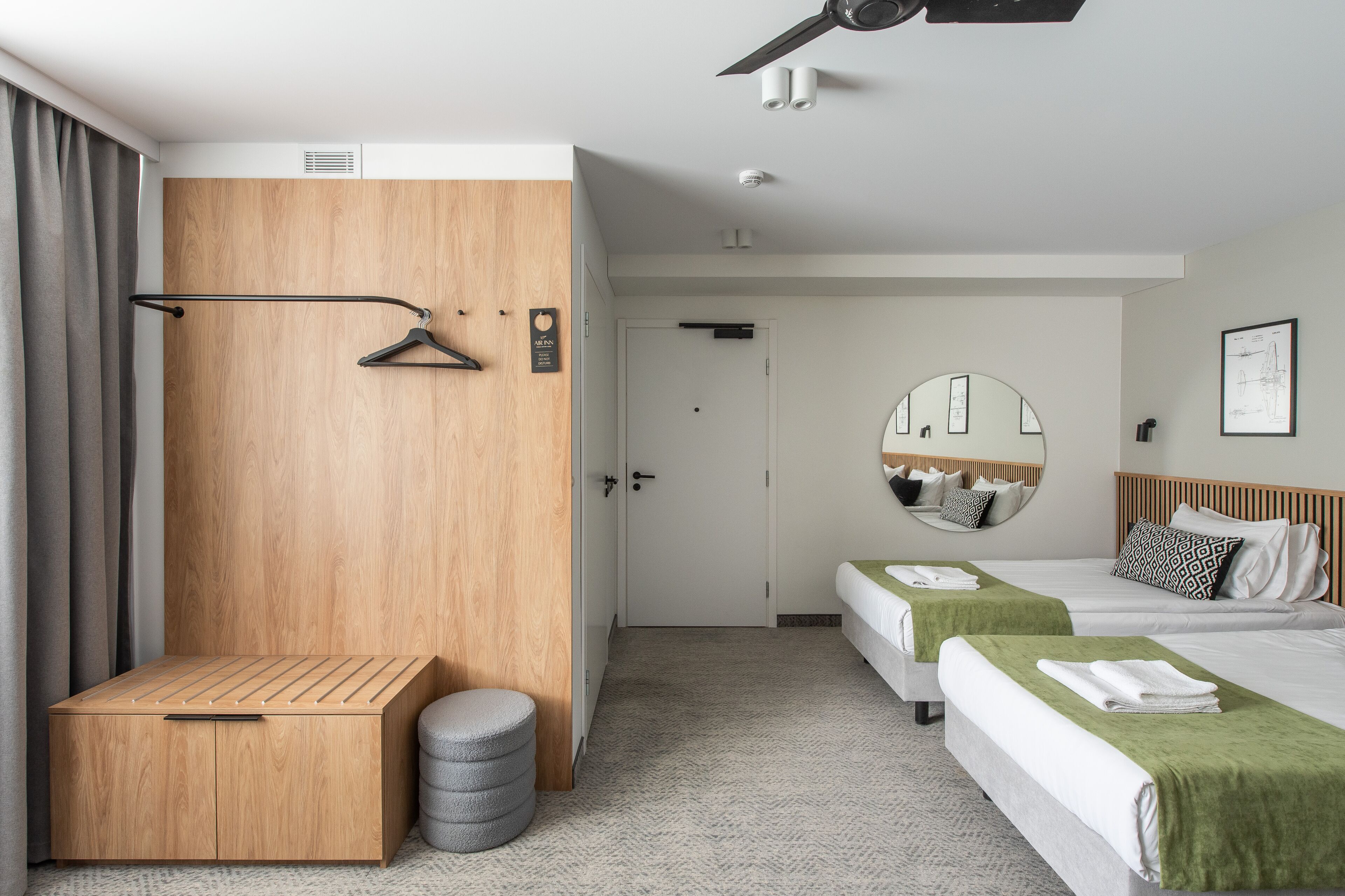 A hotel room with a wooden wardrobe, a small seating area, a mirror, two beds with green towels, and a ceiling fan.