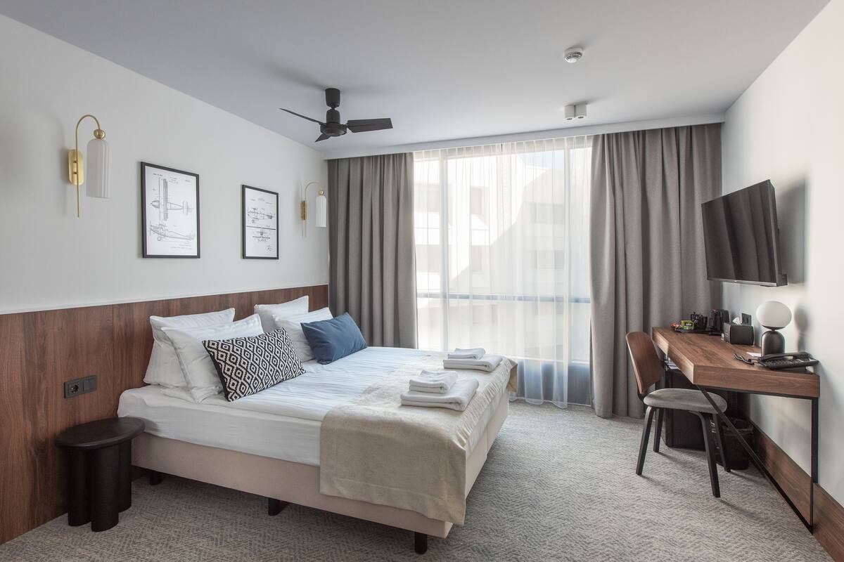 A modern hotel room with a bed, a desk, a chair, a TV, and a ceiling fan.