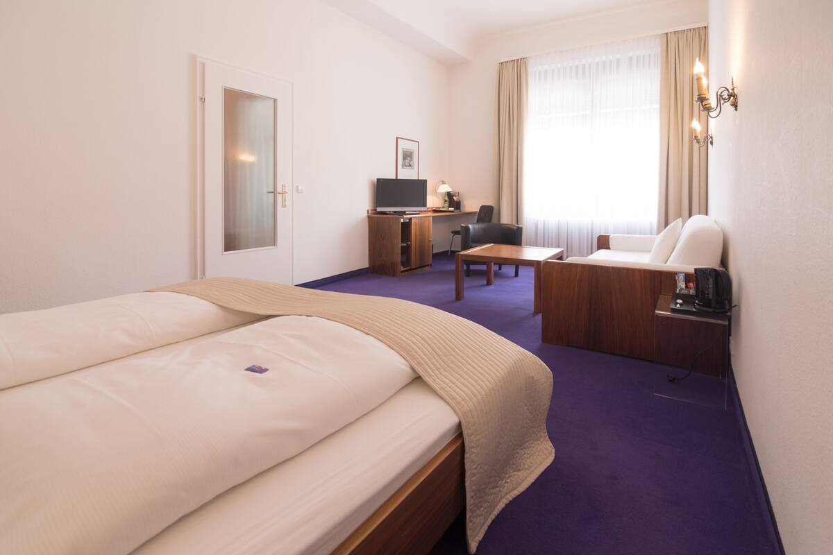 A hotel room with a large bed, a desk with a computer, a chair, a coffee maker, and a window with curtains.