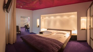 Habitación de hotel con una cama grande, un escritorio y una silla. Hay un televisor montado en la pared y un techo con un diseño rojo.