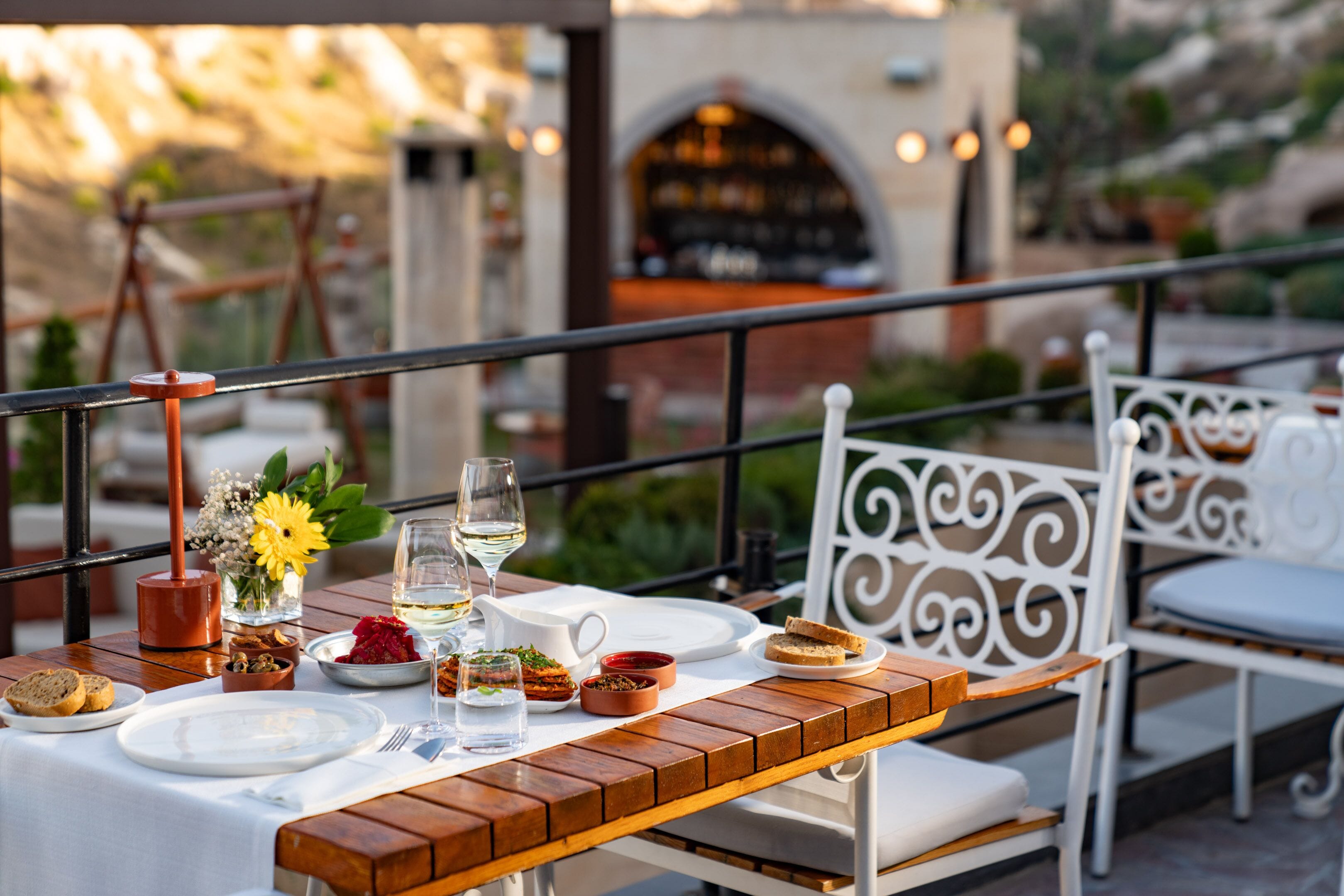 A dining table set with plates, glasses, and food, surrounded by white chairs with ornate designs, on an outdoor terrace with a railing and a building in the background.