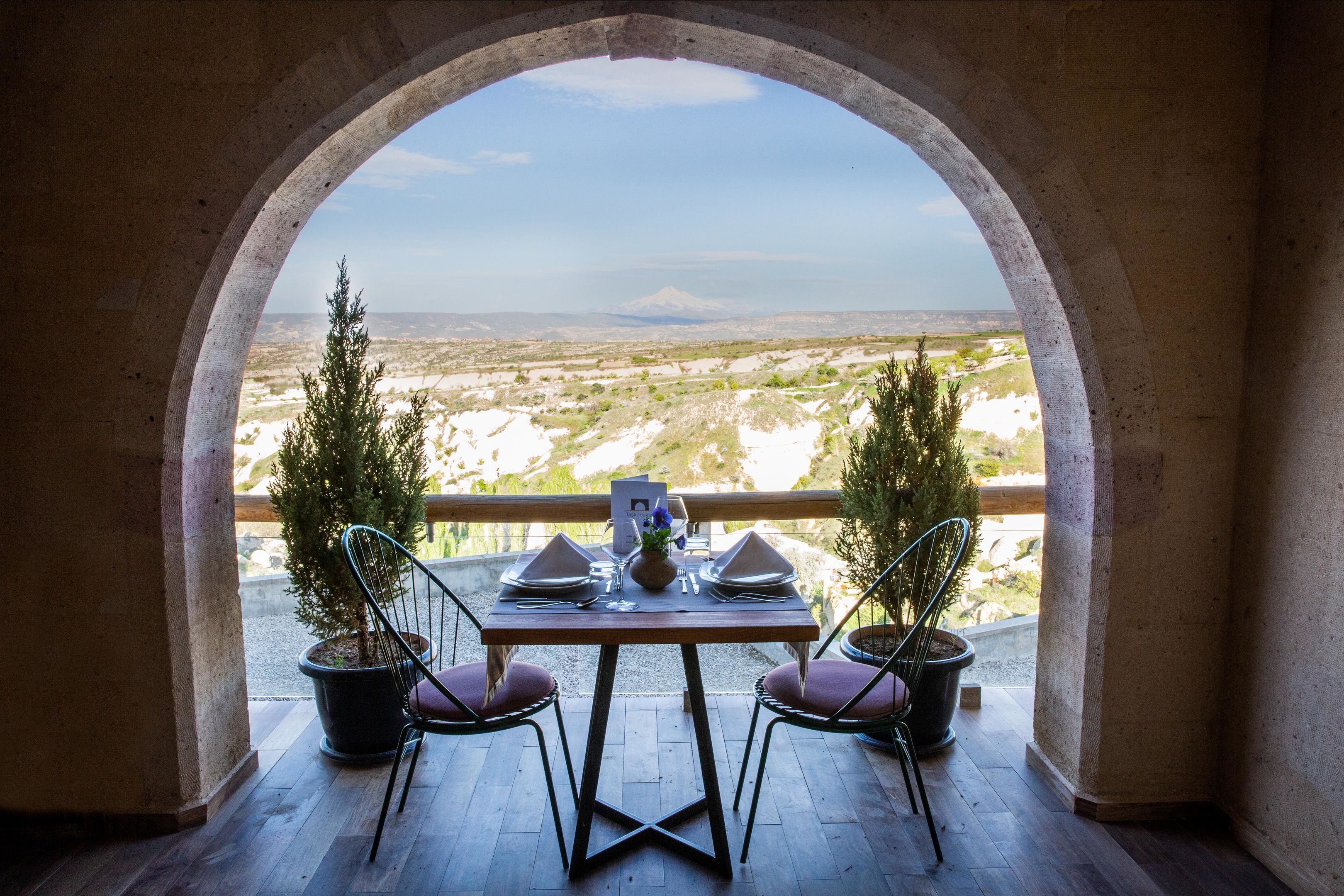 A dining table set for two with a view of a mountainous landscape through an arched window.