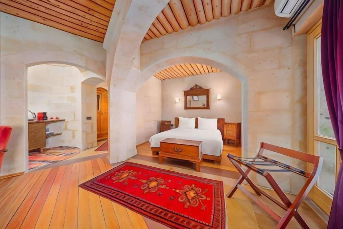 Un dormitorio con techo de madera, una cama con ropa blanca, un tapete rojo estampado y una silla de madera.