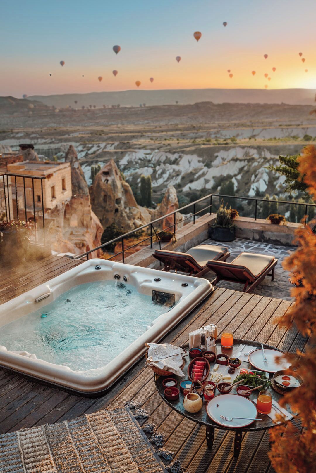 A hot tub on a wooden deck with a table set for a meal, overlooking a landscape with rock formations and hot air balloons in the distance.
