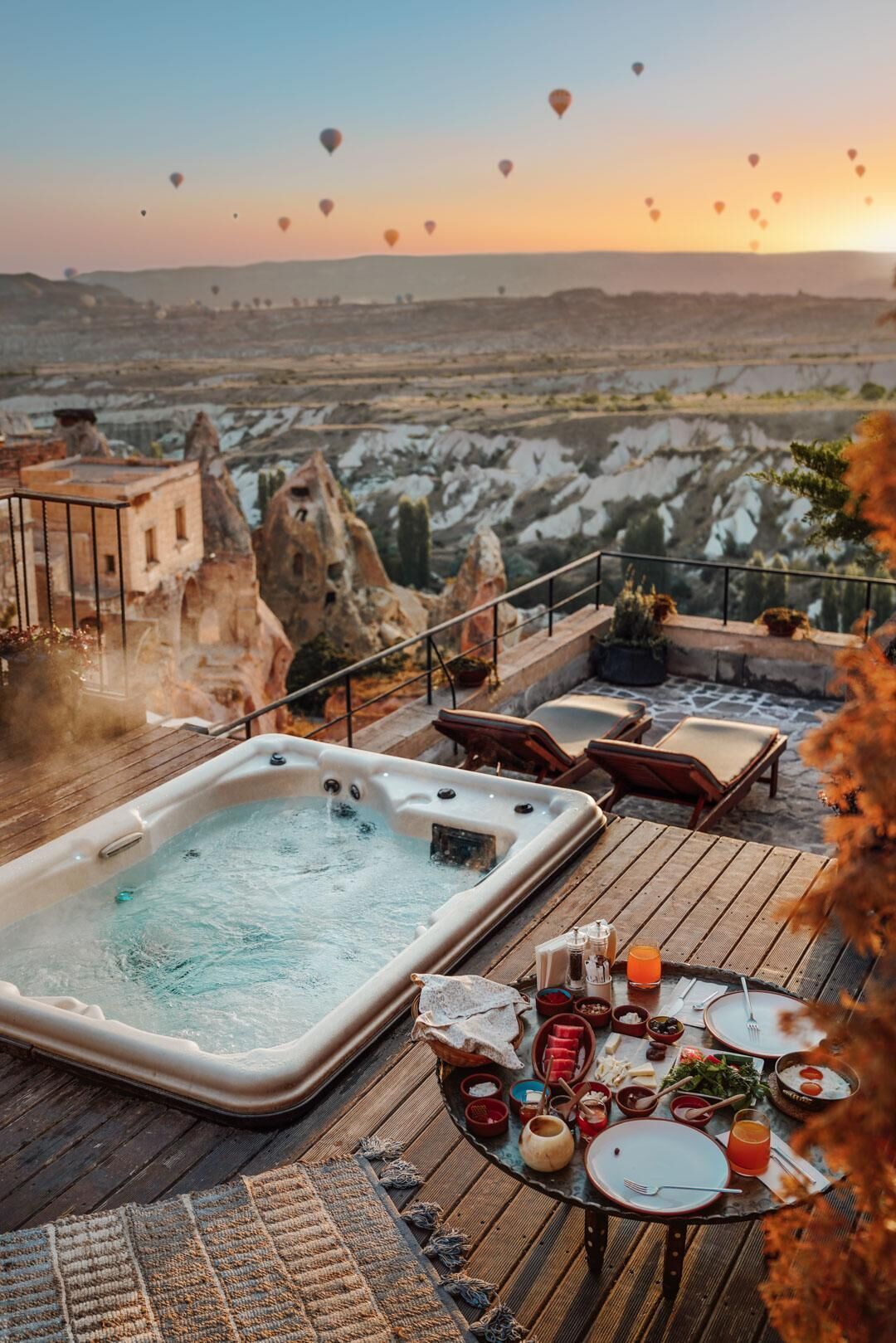 A hot tub on a wooden deck with a table set for dining, overlooking a scenic landscape with hot air balloons.