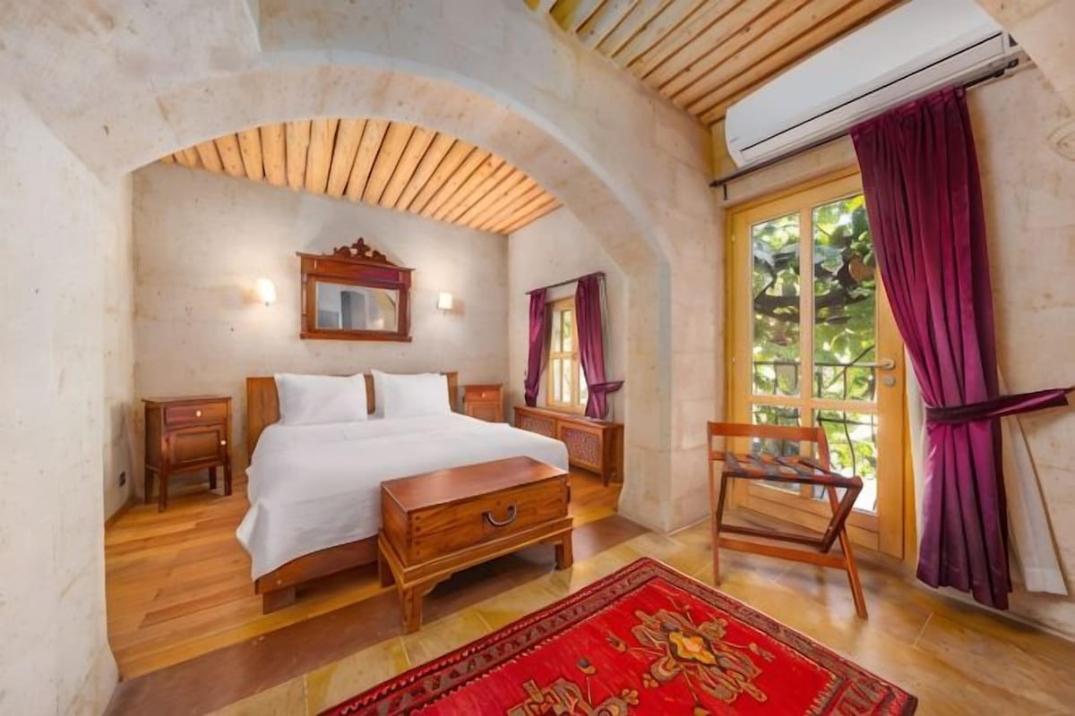 A bedroom with a bed, wooden furniture, a red rug, a window with curtains, and an arched ceiling.