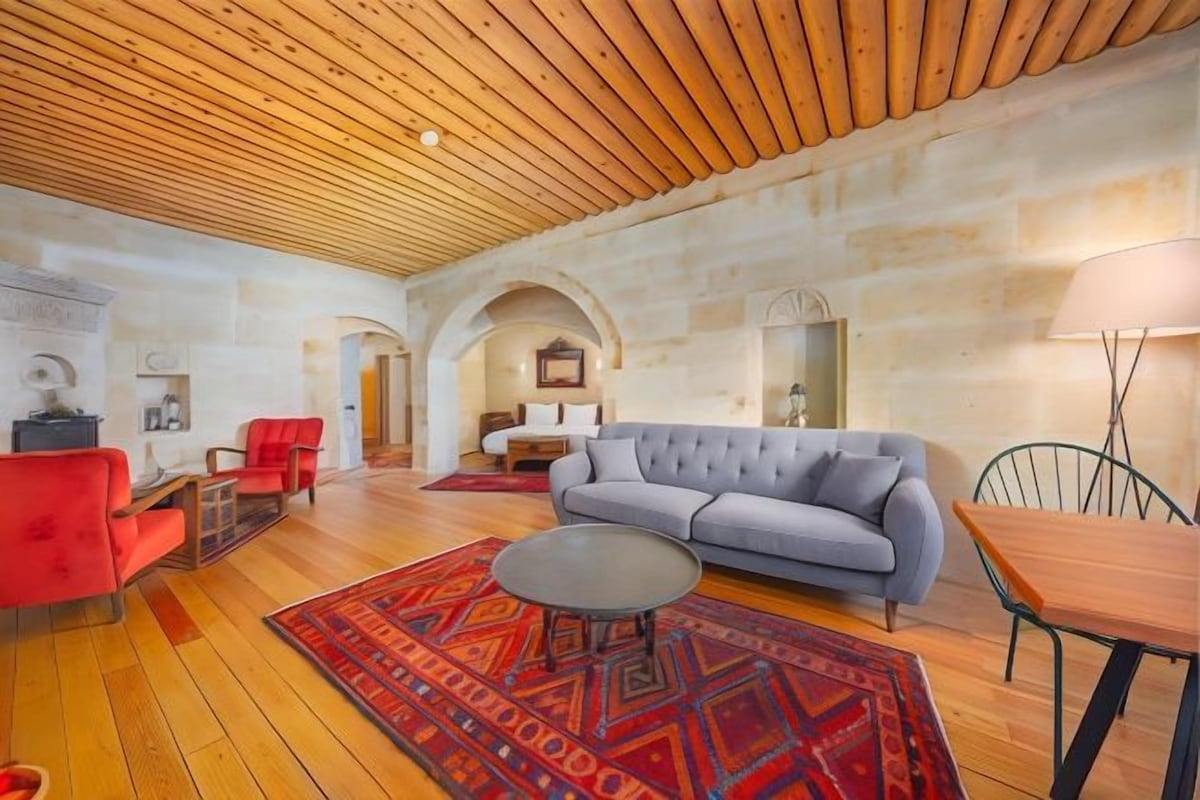 A spacious living room with a wooden ceiling, a grey sofa, red chairs, a round coffee table, and a patterned rug.