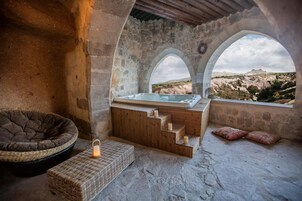 Ein Zimmer mit Steinwänden, einer Holzbank, einem kleinen Tisch mit Kerzen und Blick auf eine gebirgige Landschaft durch Rundbogenfenster.