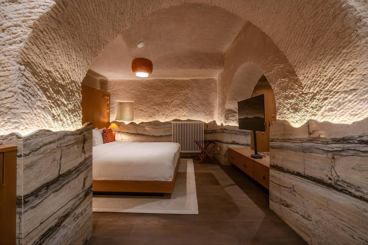 Un dormitorio con cama, mesita de noche, lámpara y televisor. La habitación tiene un techo abovedado único y paredes de piedra natural.
