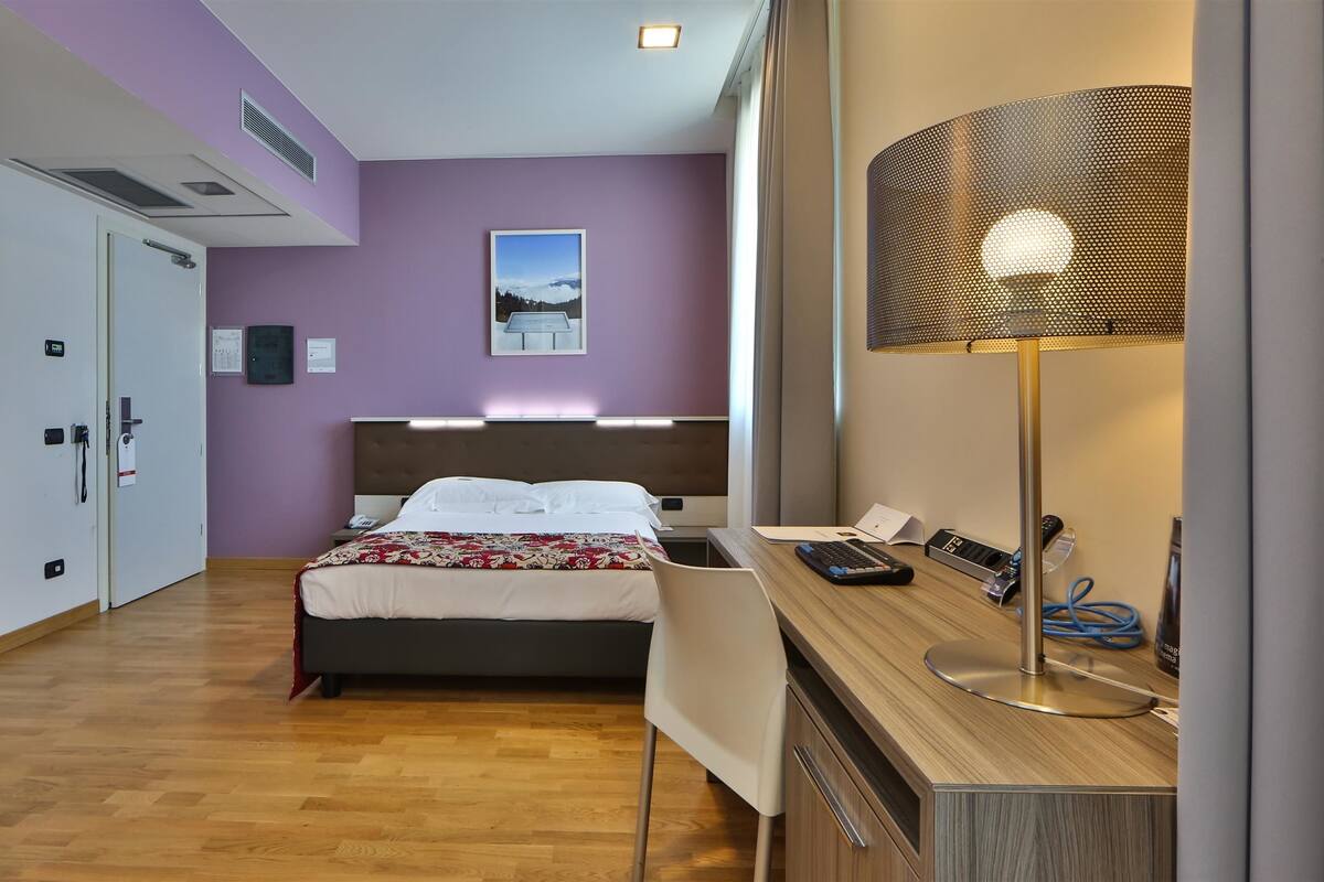 A hotel room with a bed, desk, and lamp. There is a door to another room and a window with curtains.
