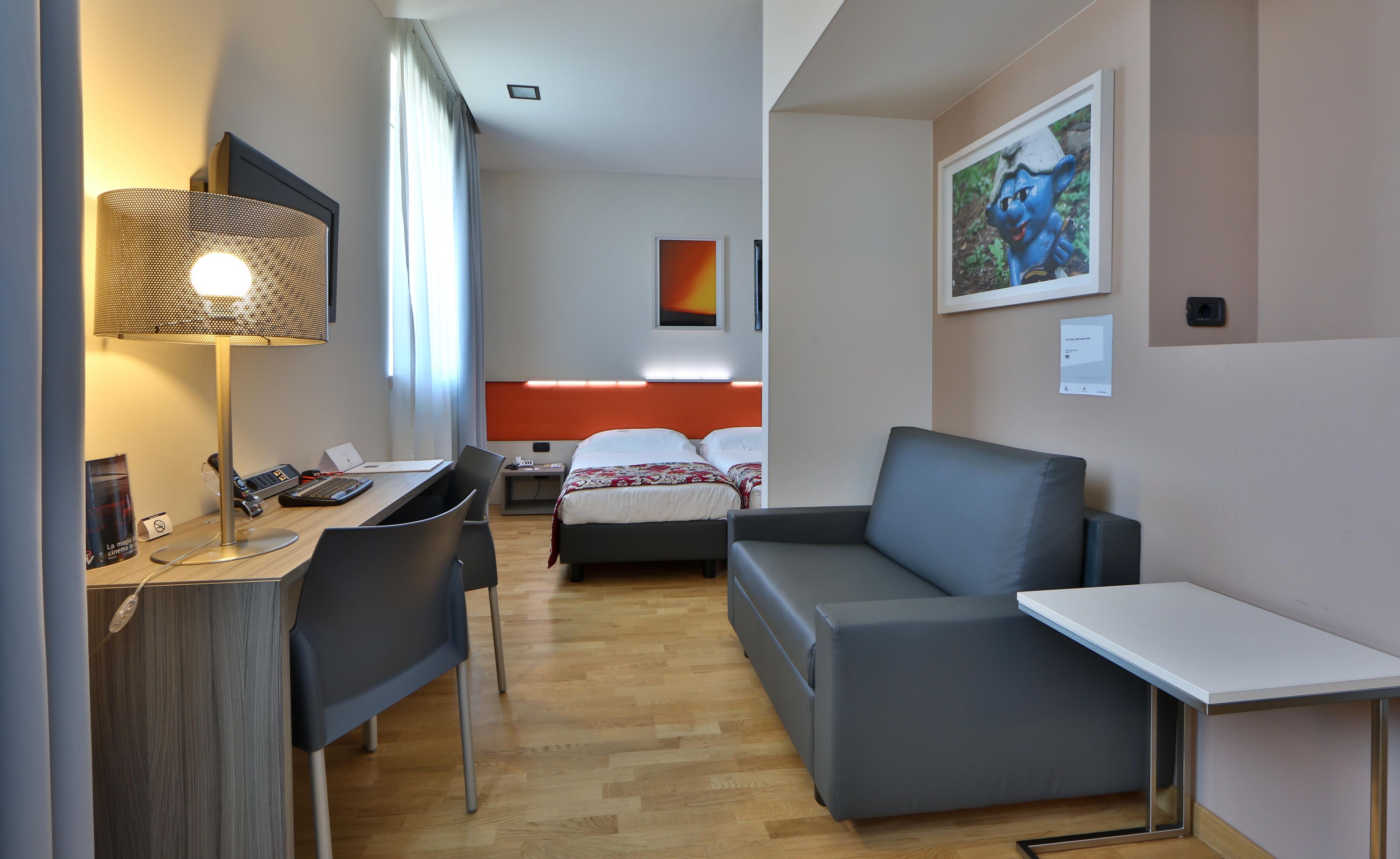 Ein Hotelzimmer mit Schreibtisch, zwei Stühlen, einer Couch und einem Bett mit rotem Kopfteil, sowie einem Bild an der Wand.