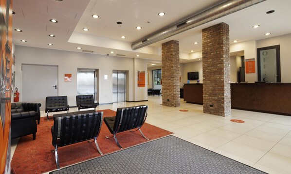 A lobby with a reception desk, two elevators, black leather chairs, a fire extinguisher, and a fire exit sign. - Best Western Falck Village Hotel (Sesto San Giovanni)