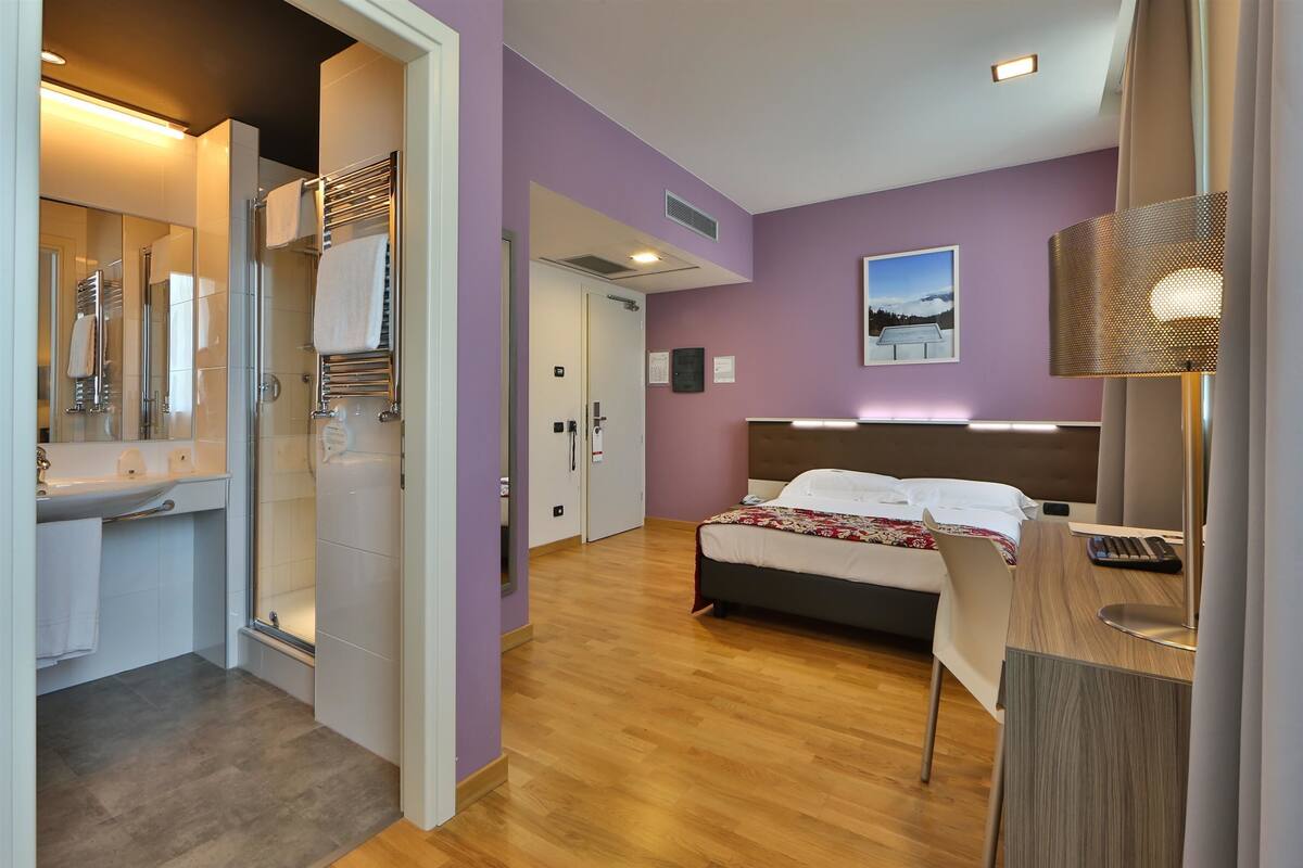 A hotel room with a bed, a desk, a bathroom with a shower, and a mirror.