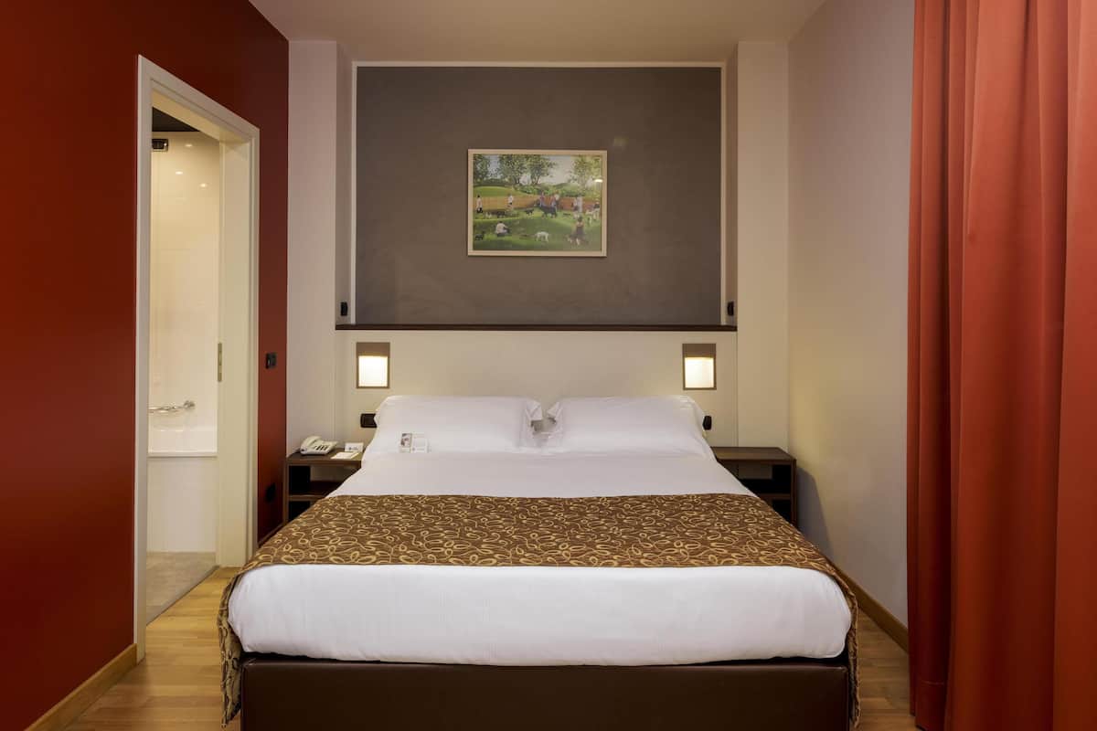 A hotel room with a large bed, a painting on the wall, a bedside table with a lamp, and a door leading to a bathroom.