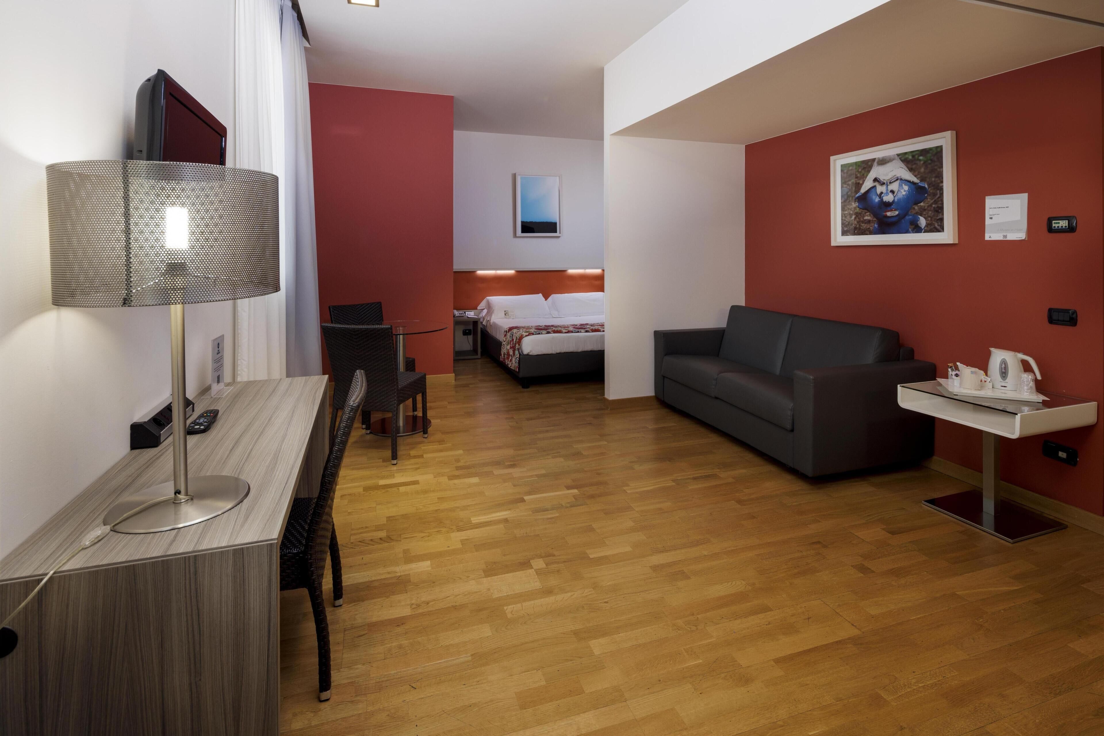 Hotellihuone, jossa on puulattia, kirjoituspöytä lampun kanssa, harmaa sohva, valkoisella päätyosalla varustettu sänky ja taulu seinällä.