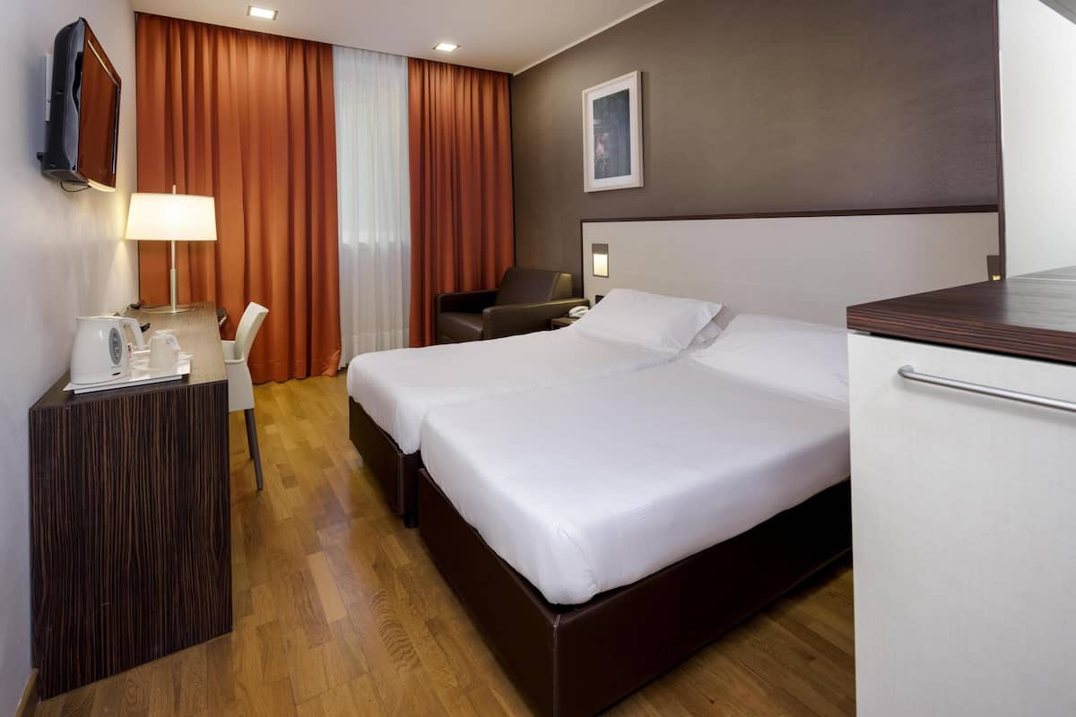 A hotel room with a large bed, a desk with a lamp, a chair, a TV mounted on the wall, and a picture on the wall.
