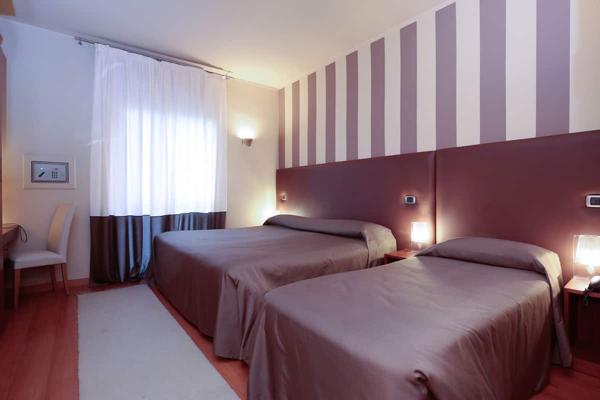 A hotel room with two beds, a desk, a chair, a window with curtains, and a striped wall.