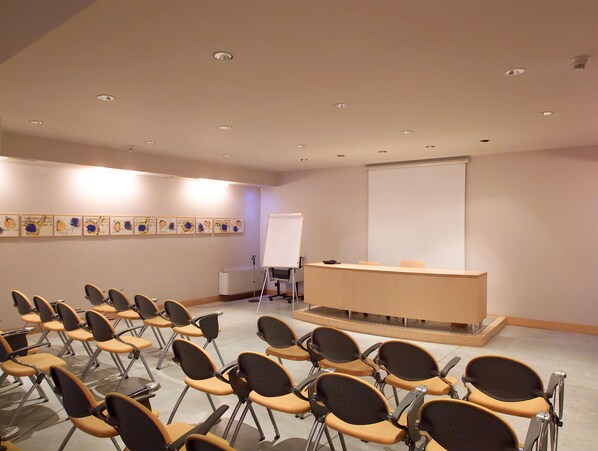 A conference room with rows of chairs, a podium, a projector screen, and artwork on the walls. - Hotel Aleramo (Asti)