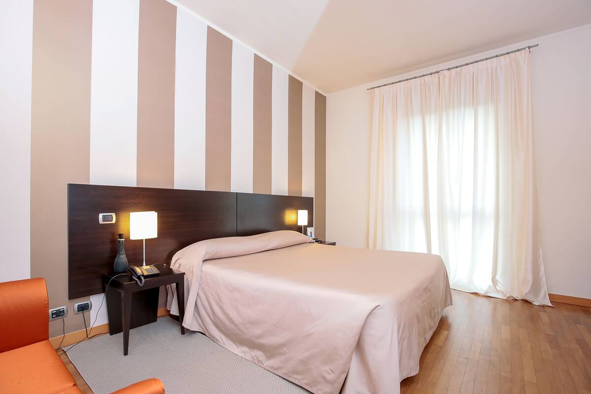 A hotel room with a large bed, a nightstand with a lamp, a chair, and a window with curtains.
