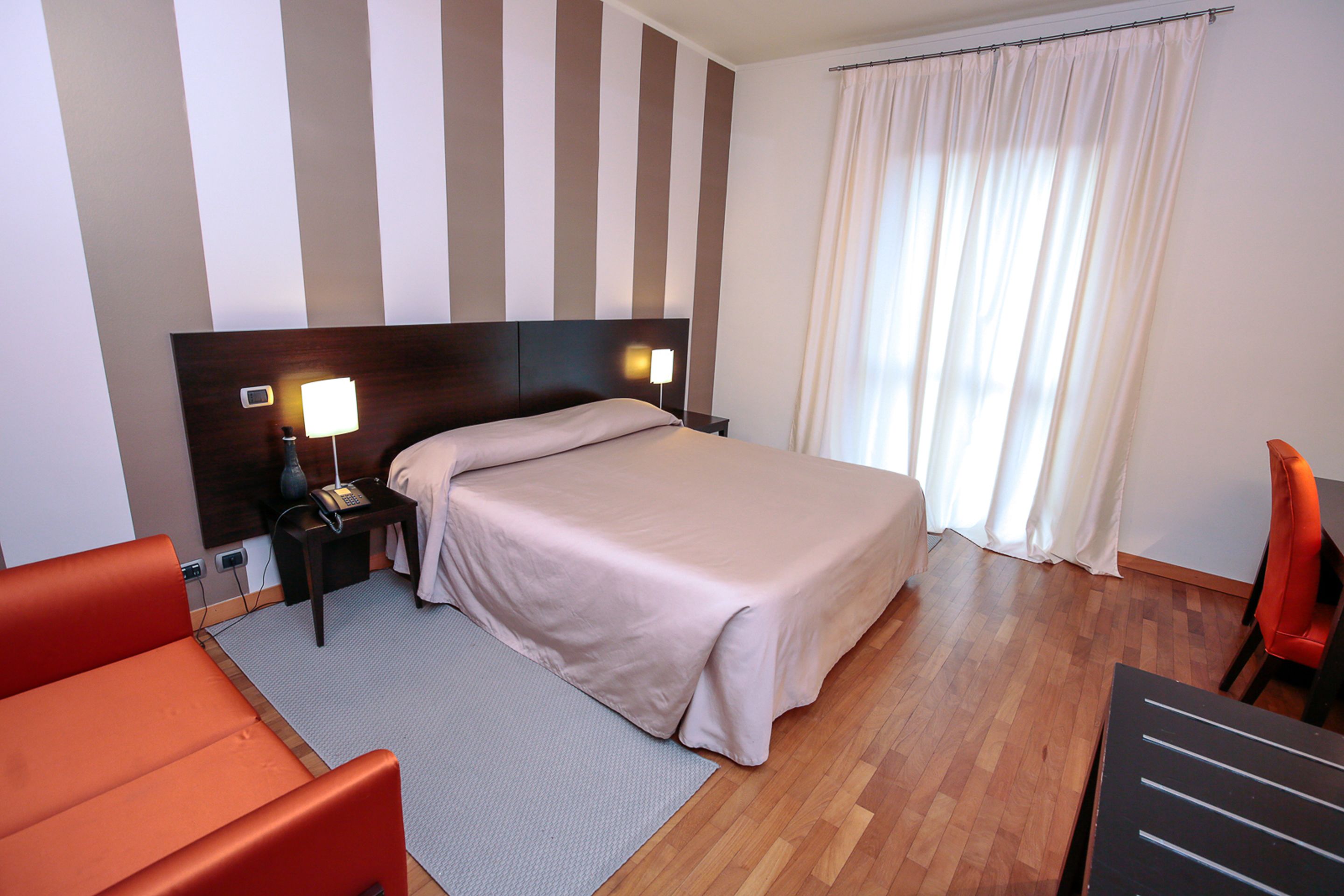 Une chambre d’hôtel avec un lit, une table de chevet, une lampe, une chaise et une fenêtre avec des rideaux.