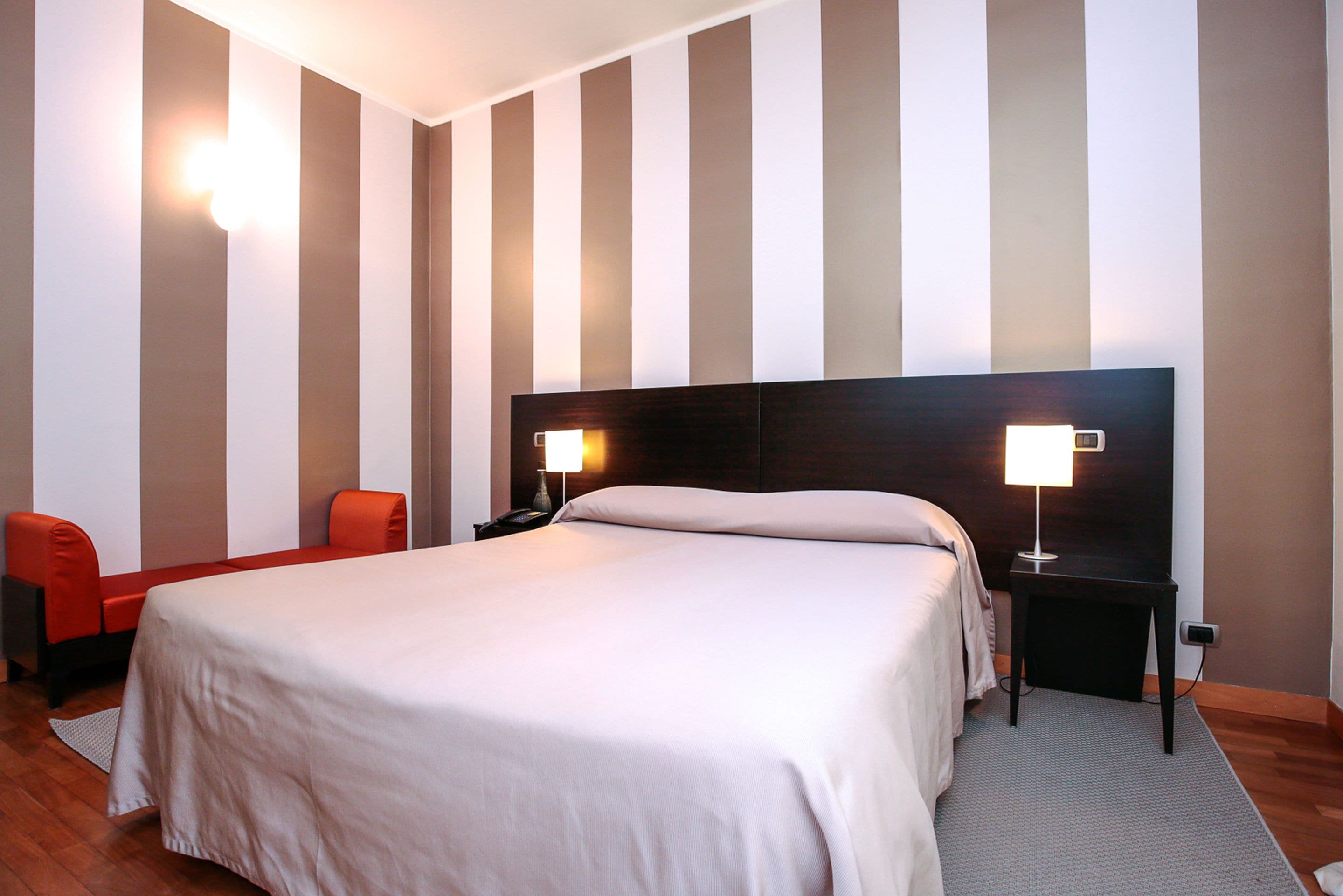 Una habitación de hotel con una cama grande, dos mesitas de noche con lámparas, una silla roja y una pared a rayas.