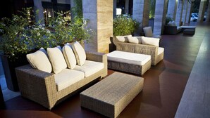 A wicker outdoor seating area with cushions and a bench, surrounded by greenery and a tiled floor. - Hotel Aleramo (Asti)