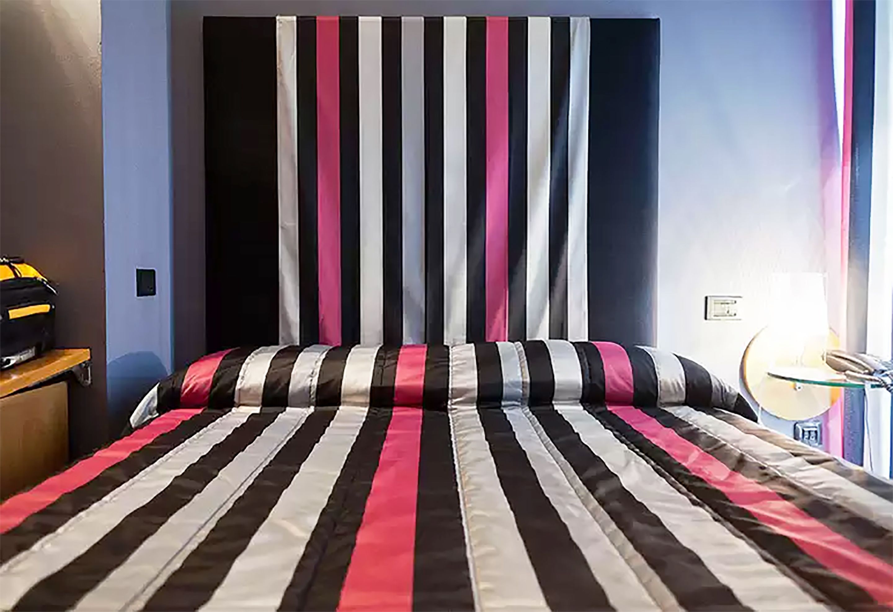 Una cama con una colcha a rayas, una mesita de noche con una lámpara y una ventana con cortinas.