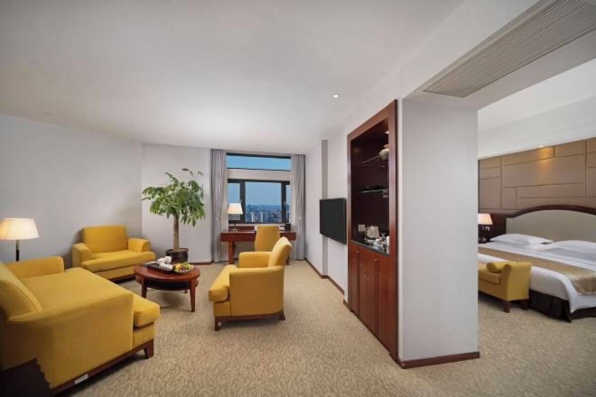 Habitación de hotel con una cama, dos sillones amarillos, una mesita con una lámpara, un televisor y un ventanal que ofrece vistas a la ciudad.