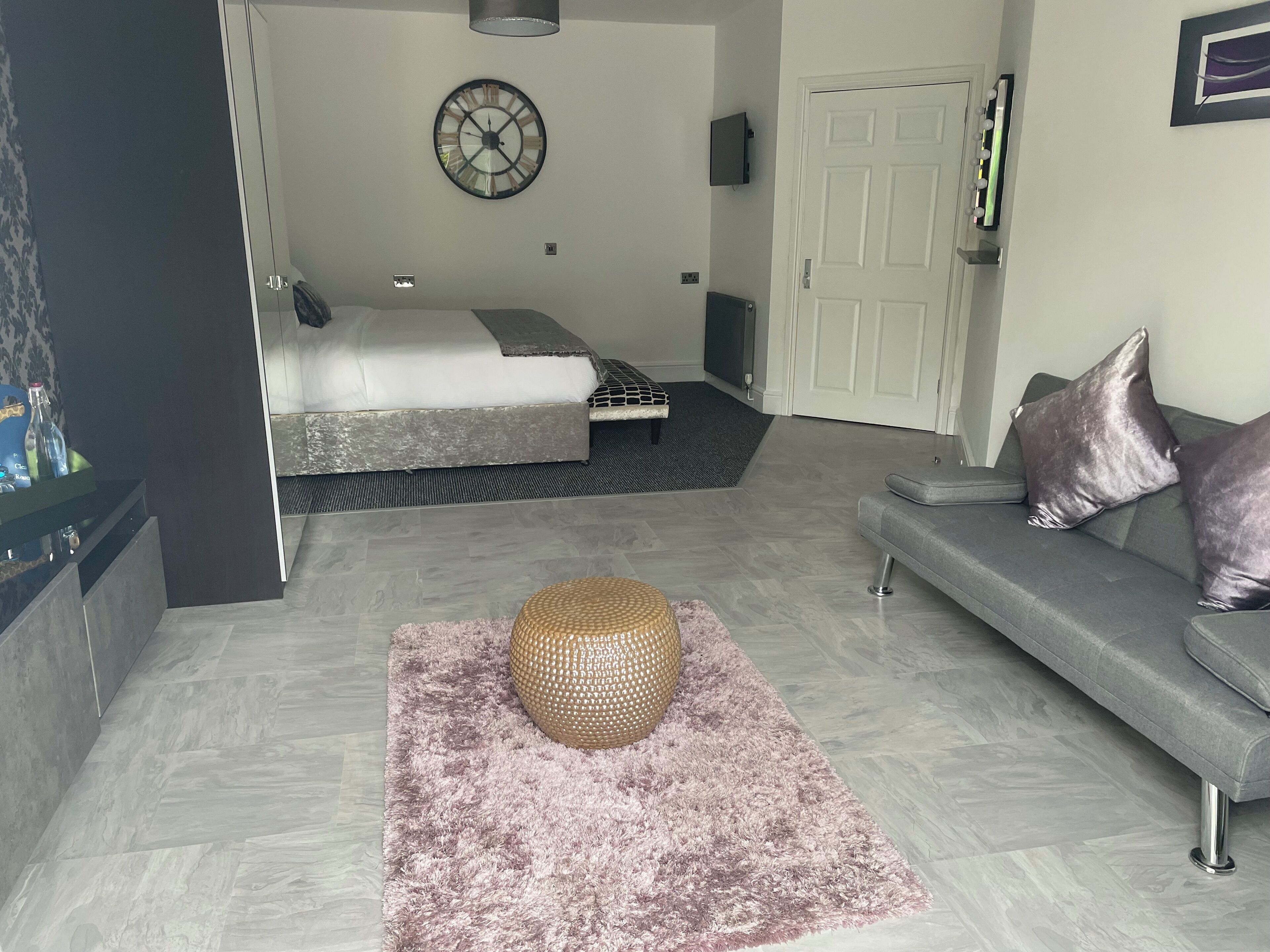 Een moderne woonkamer met een grijze bank, een rond geweven poef en een bed met een grijs hoofdeinde.