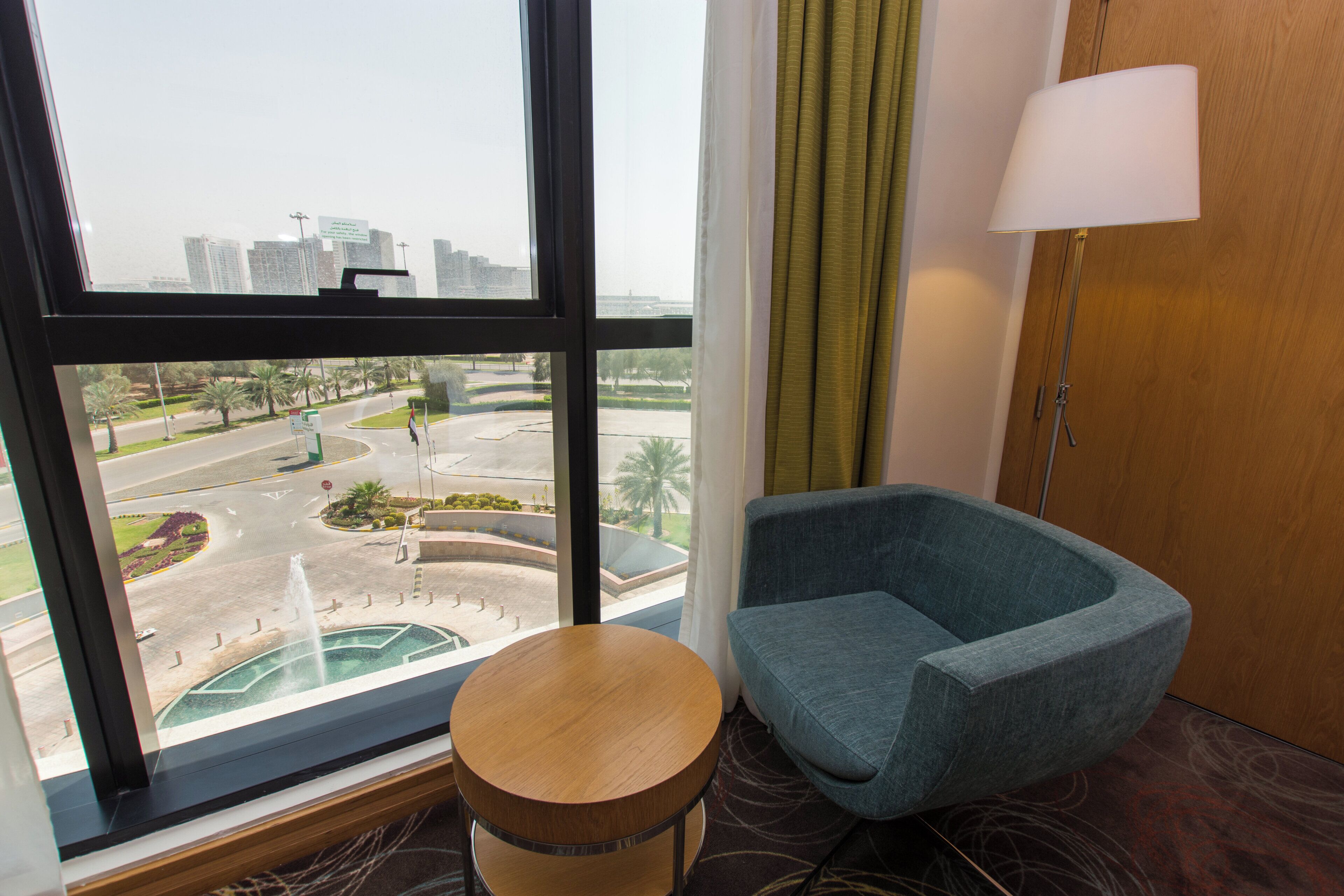 A hotel room with a view of a cityscape, a round wooden stool, a blue armchair, and a lamp.