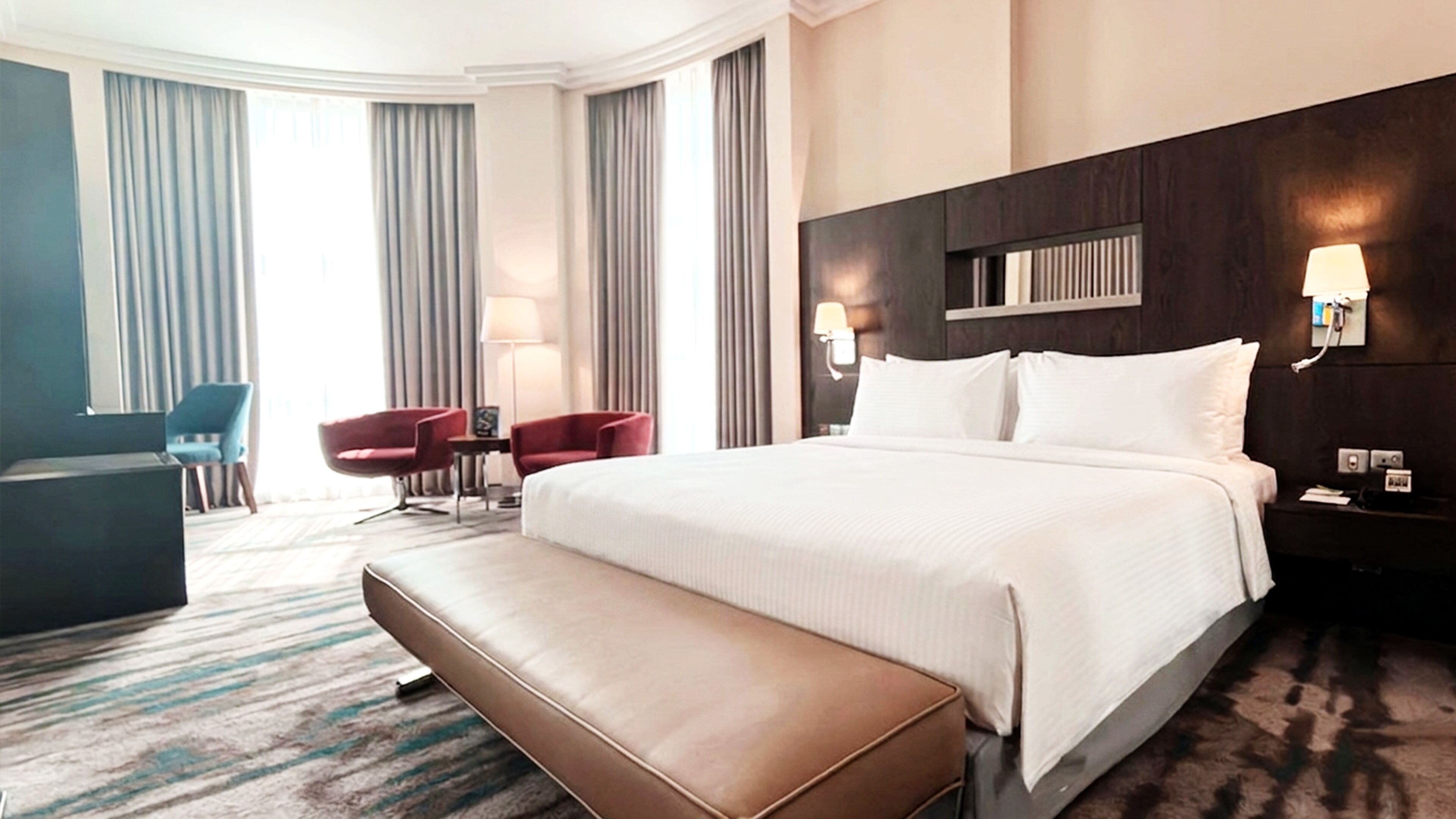 A hotel room with a large bed, a desk, two chairs, a bench, and a nightstand with a lamp.